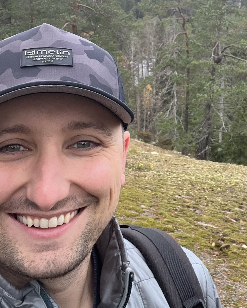 Ben Anderholm, founder of Anderholm Digital, smiling outdoors in a ball cap with a forested lake overlook behind him.